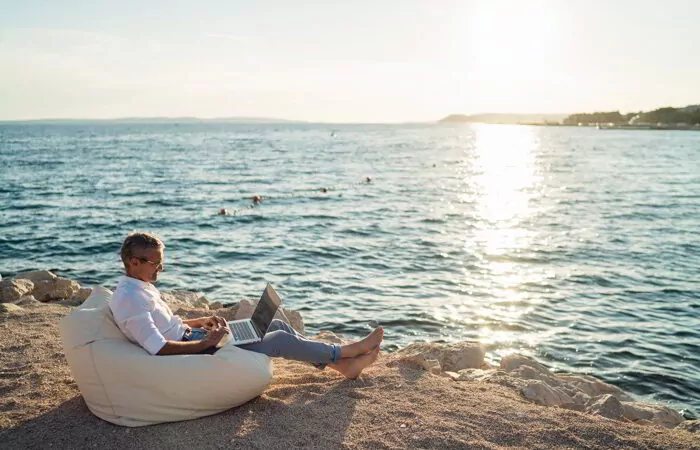 Senior man working on his laptop lying on deck chair on the beach during sunset