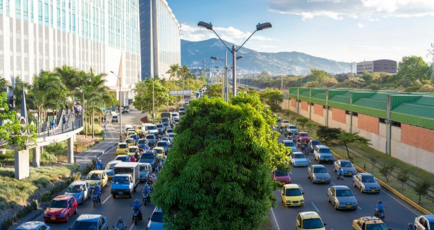 The cityscape of El Poblado, Medellin, Colombia, with traffic spanning 6 lanes divided by trees in the middle and mountains off in the distance.
