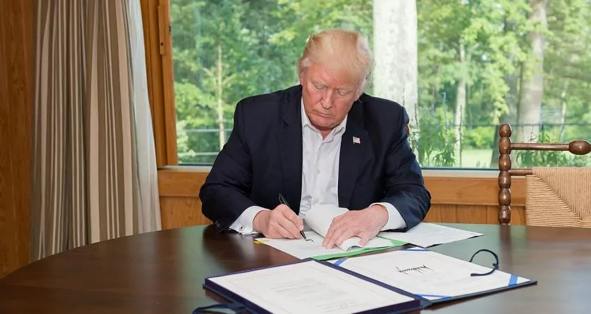 Donald Trump Signing A Bill In Front Of A Window. The View Outside Is Of Green Trees, Trump Is Sitting At A Wooden Table And Not Wearing A Tie