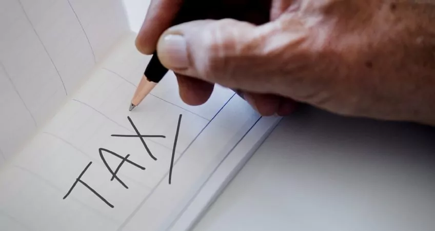hand holding a pencil which has written the word tax in capital letters on a piece of paper
