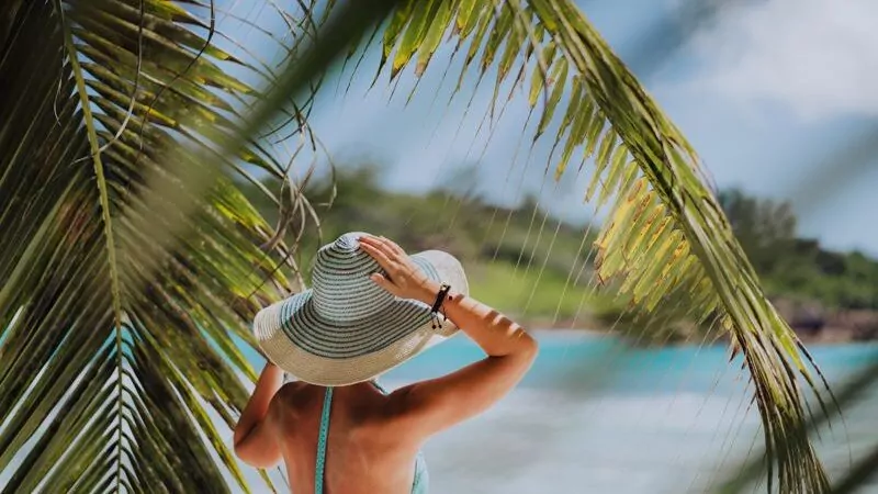 Woman on the beach in the palm trees shadow wearing blue hat.