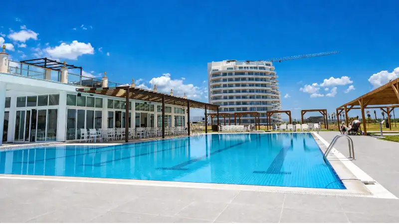 Bright outdoor swimming pool at a modern luxury resort, with a contemporary hotel building, poolside lounge areas, and a vivid blue sky.