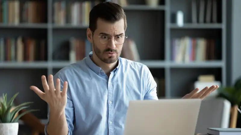 Unhappy young Caucasian male worker in glasses look at laptop screen shocked by gadget breakdown or operational problems. Frustrated man confused surprised by unexpected error on computer device.